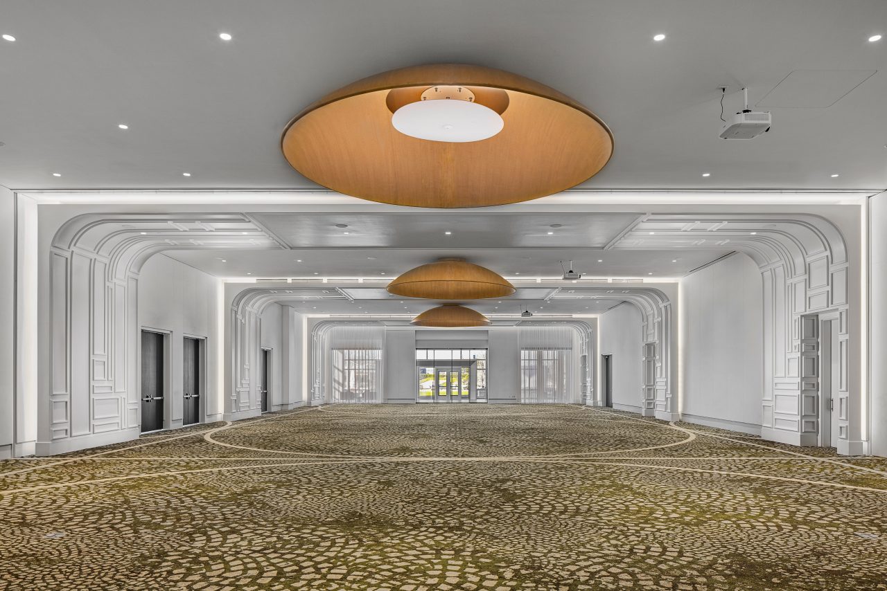 The Dream Ballroom features a spacious, elegant hallway with patterned carpet, large round ceiling lights, and ornate white molding. Multiple doors line the sides, leading to a distant entrance. Ideal for event venues or ballroom rentals, it promises an enchanting experience.