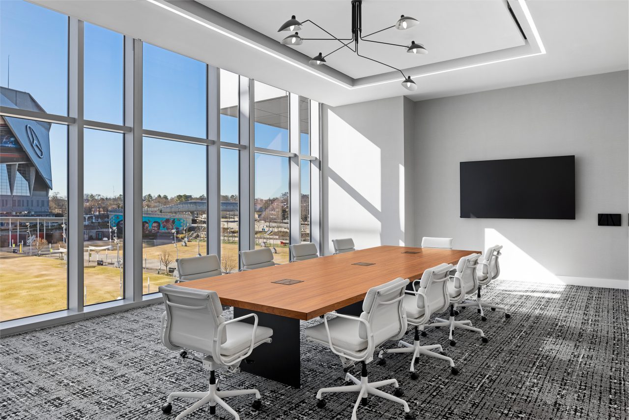 A modern conference room features a long wooden table, white chairs, and a large TV screen on the wall. Floor-to-ceiling windows offer sweeping views of the cityscape, reminiscent of Mt. Vernon's charm. Perfectly suited for boardrooms seeking both style and functionality.