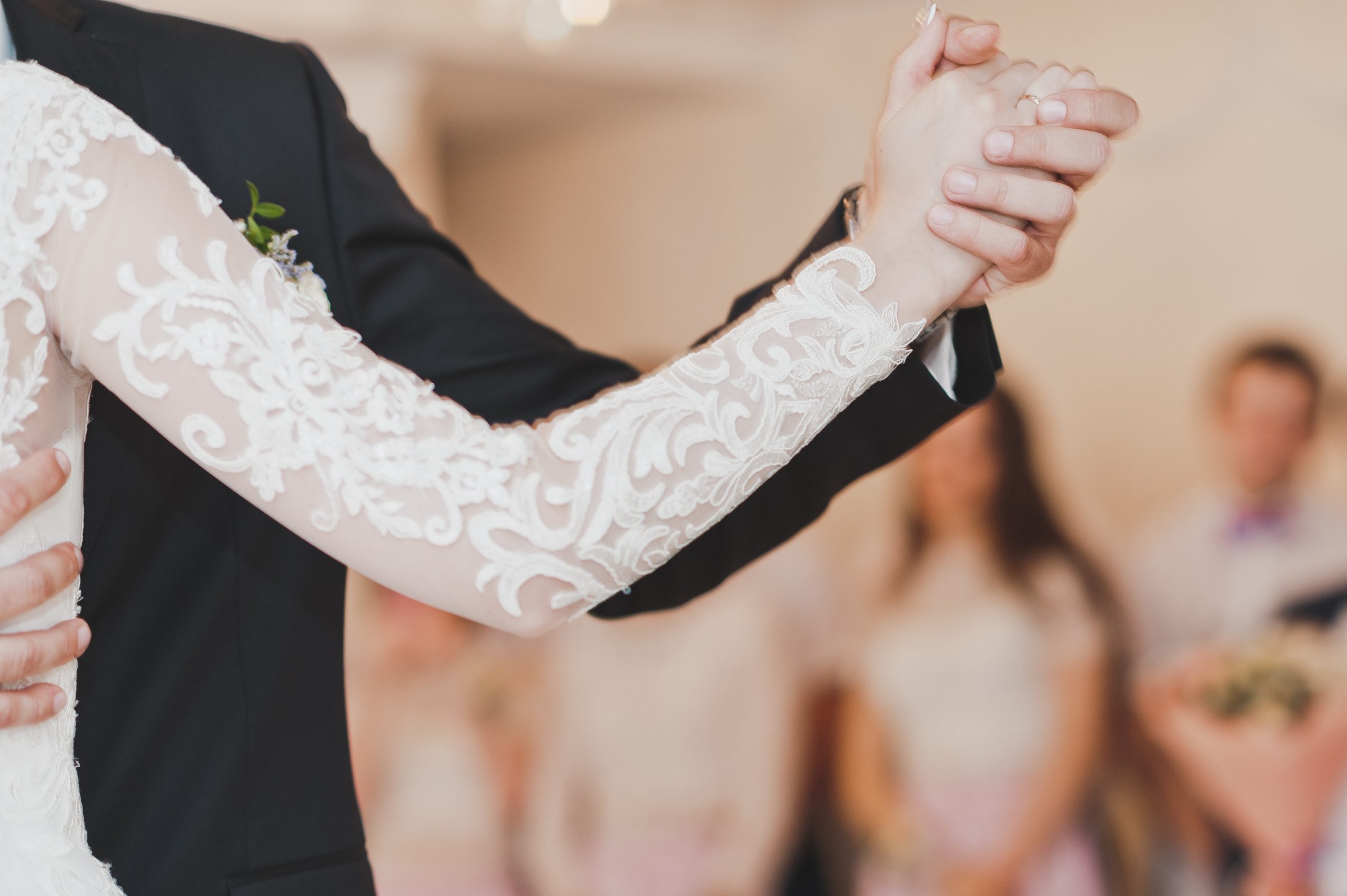 A bride and groom hold hands, gracefully moving to the rhythm they learned in dancing lessons. The bride's dress boasts lace sleeves, while the groom looks sharp in a black suit. Blurred guests mingle in the Dream Ballroom's elegant setting.