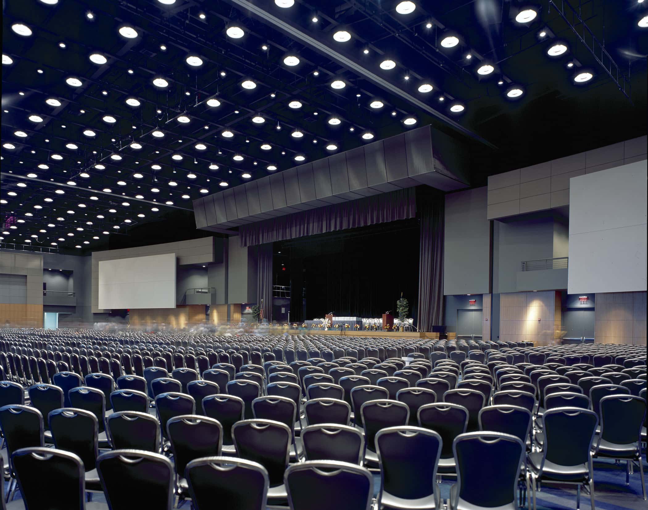 Shot from the back of a ballroom setup theater-style with blue chairs facing a stage with screens and overhead lighting