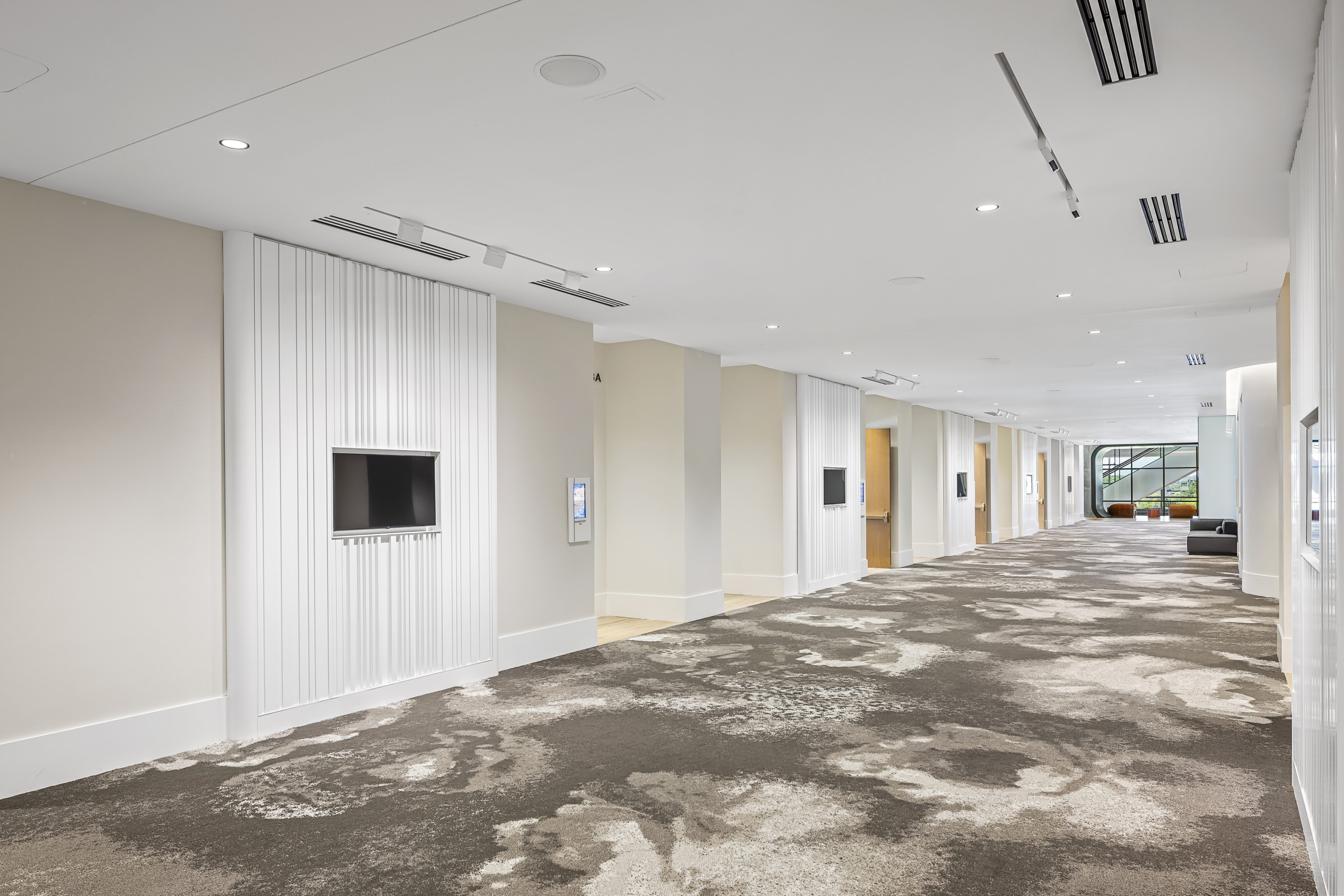 A modern, well-lit hallway with patterned carpet, white walls, recessed lighting, and wall screens leads to magnificent meeting rooms through a glass doorway at the end.