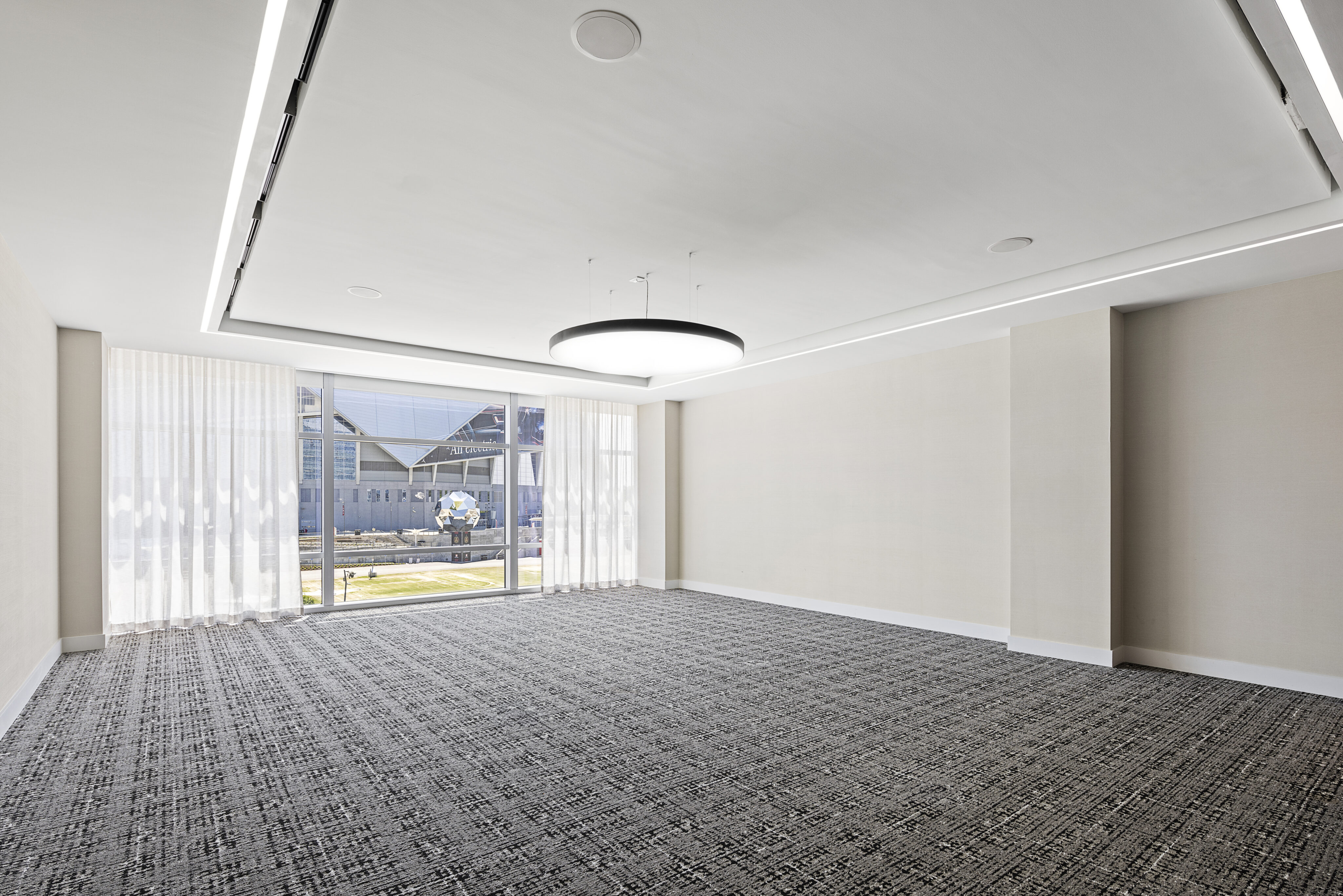 The magnificent meeting space boasts a patterned carpet, large window with sheer curtains, and a circular ceiling light. Enjoy the bright outdoor view of a stadium and lush greenery through the window.