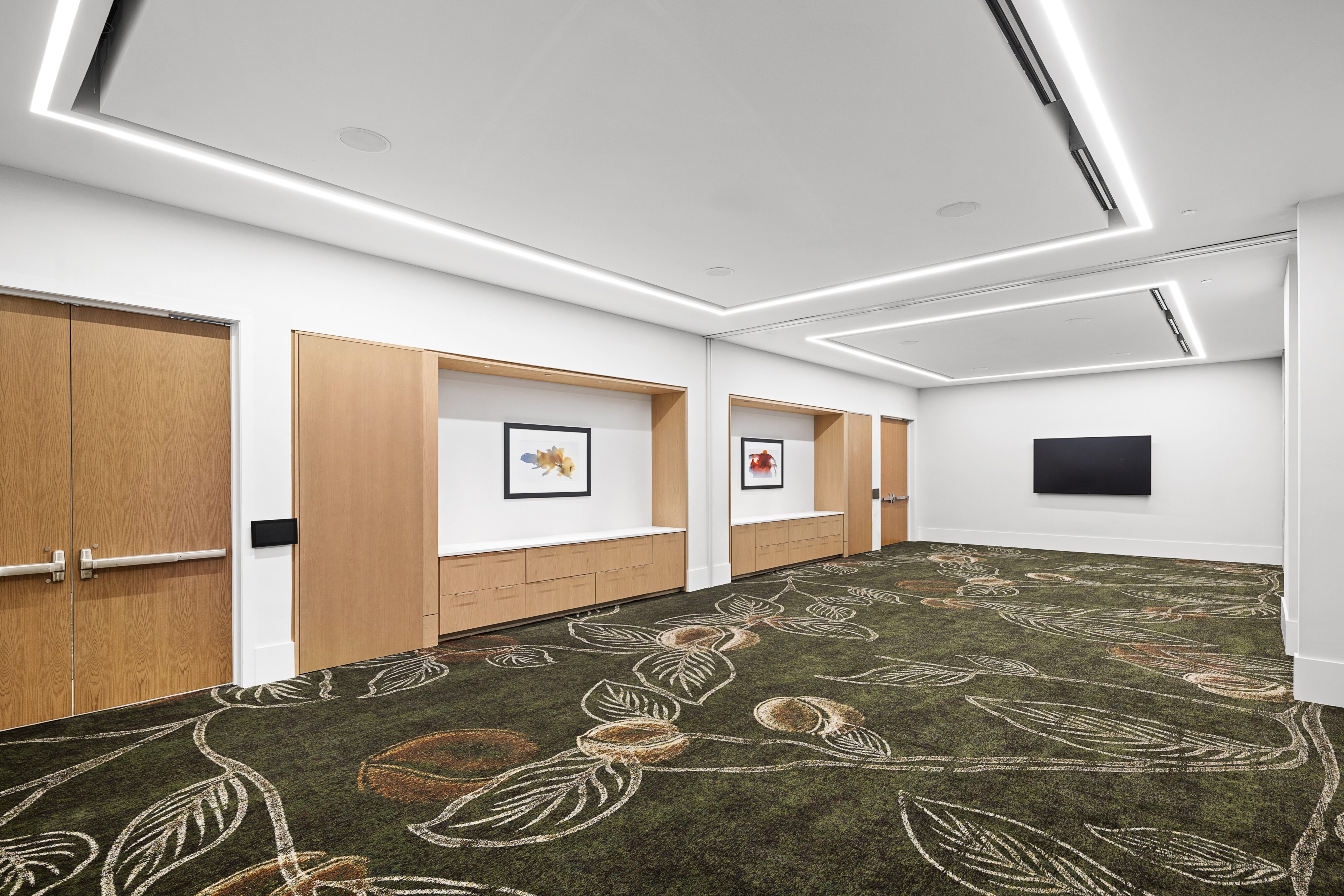 The serene meeting room features a patterned carpet, two wooden doors, and framed artworks on a beige wall. A wall-mounted black screen complements the setting, while ceiling lights softly illuminate the peaceful space.