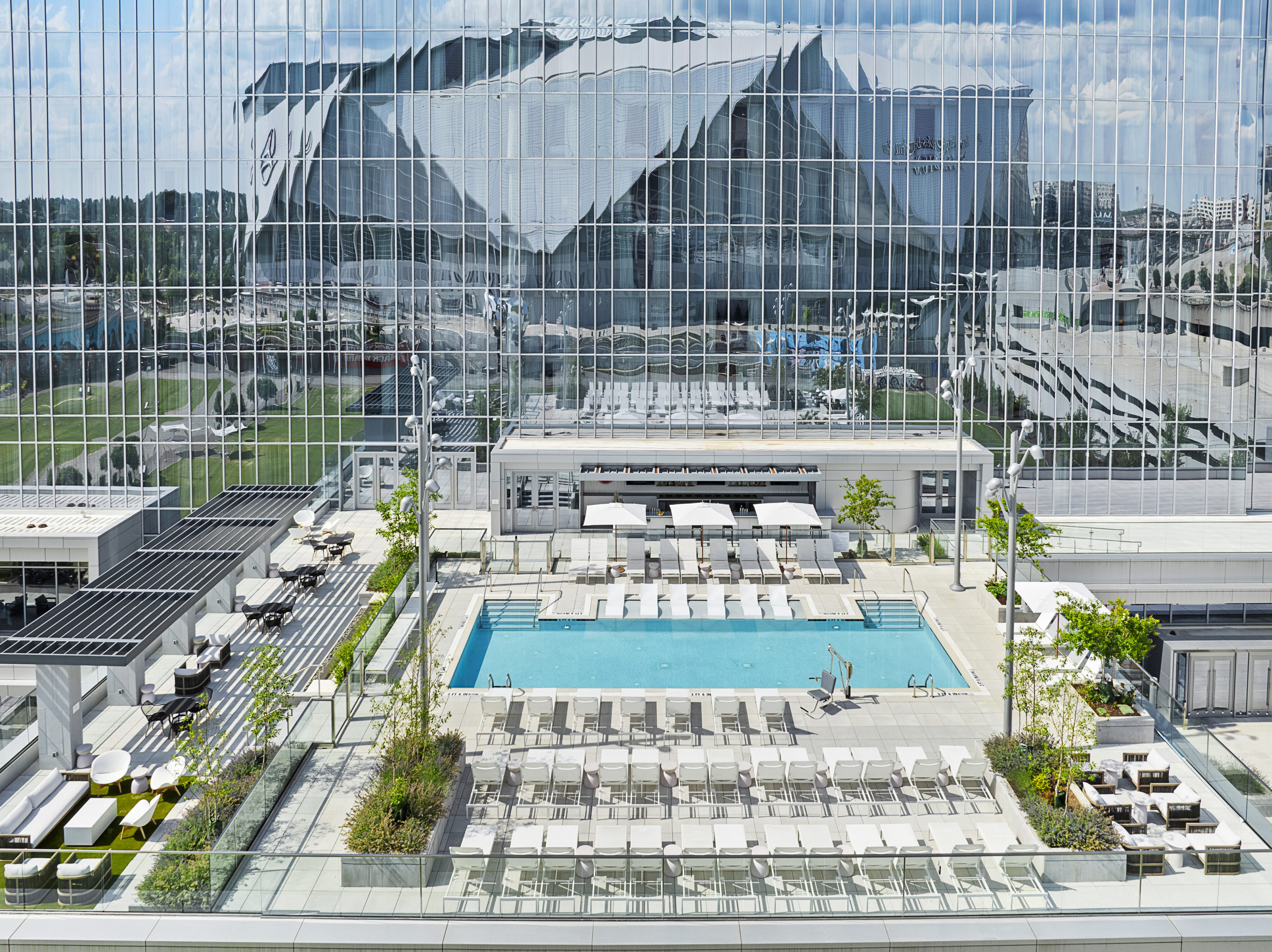Rooftop pool area with numerous lounge chairs, greenery, and a view of a modern glass building reflecting its surroundings.
