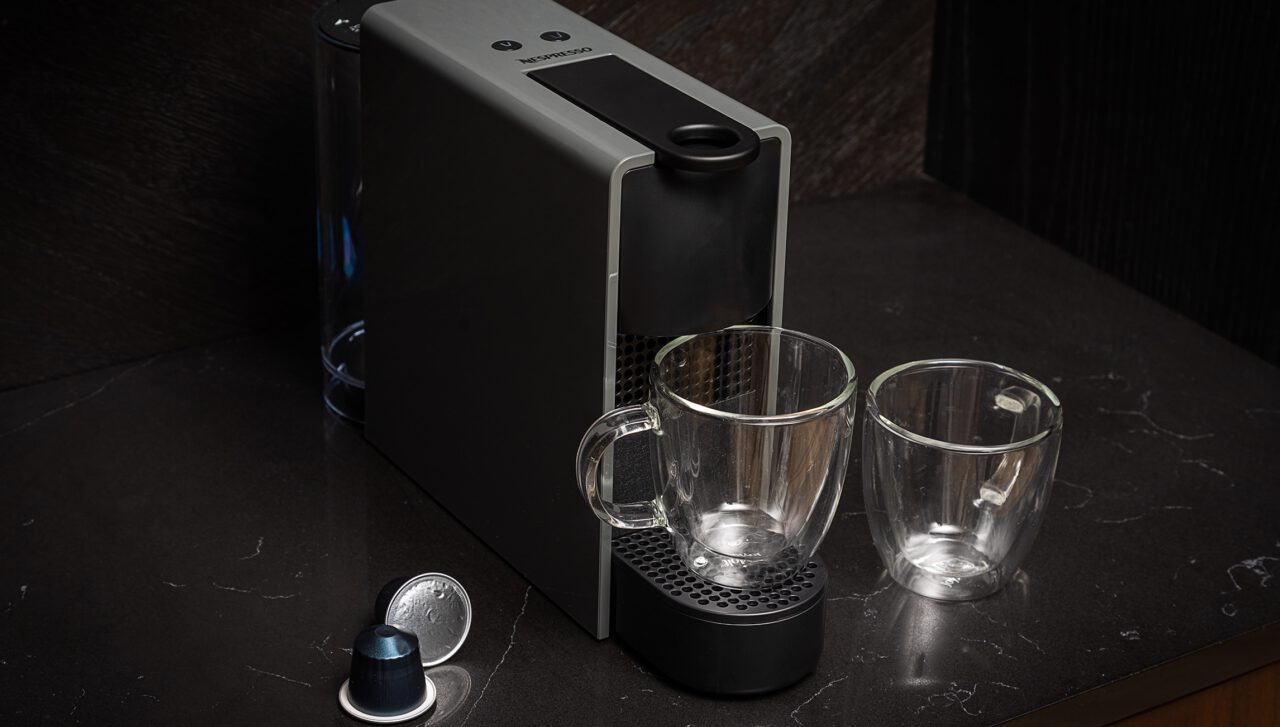 A Nespresso machine on a dark countertop with two empty glass mugs placed on the drip tray, and two coffee capsules nearby.