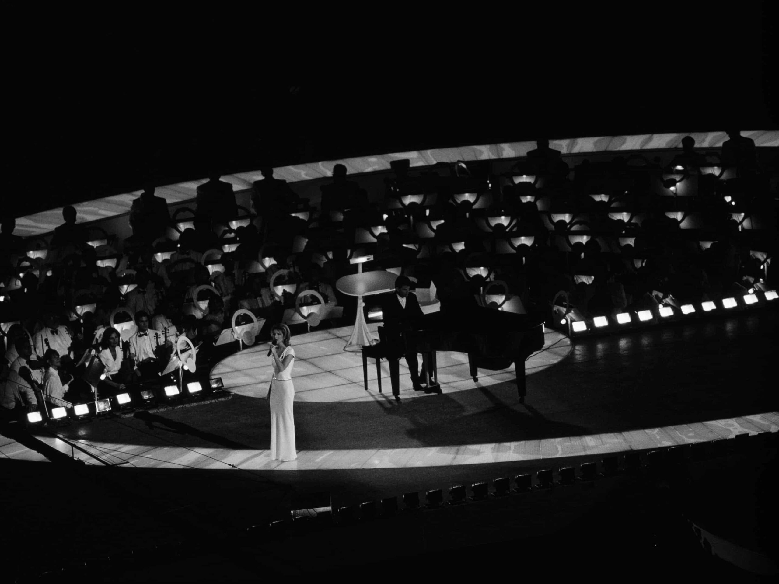 In a stunning performance, the singer in a white dress captivates on a circular stage with a grand piano and pianist. An orchestra provides a lush backdrop, reminiscent of Type II Meeting Rooms, where every detail enhances the atmosphere. The stage glows against the dark surroundings.