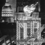 Black and white image of a lit Olympic cauldron on a metal structure, set against a cityscape with Type I Meeting Rooms and a domed building in the background.