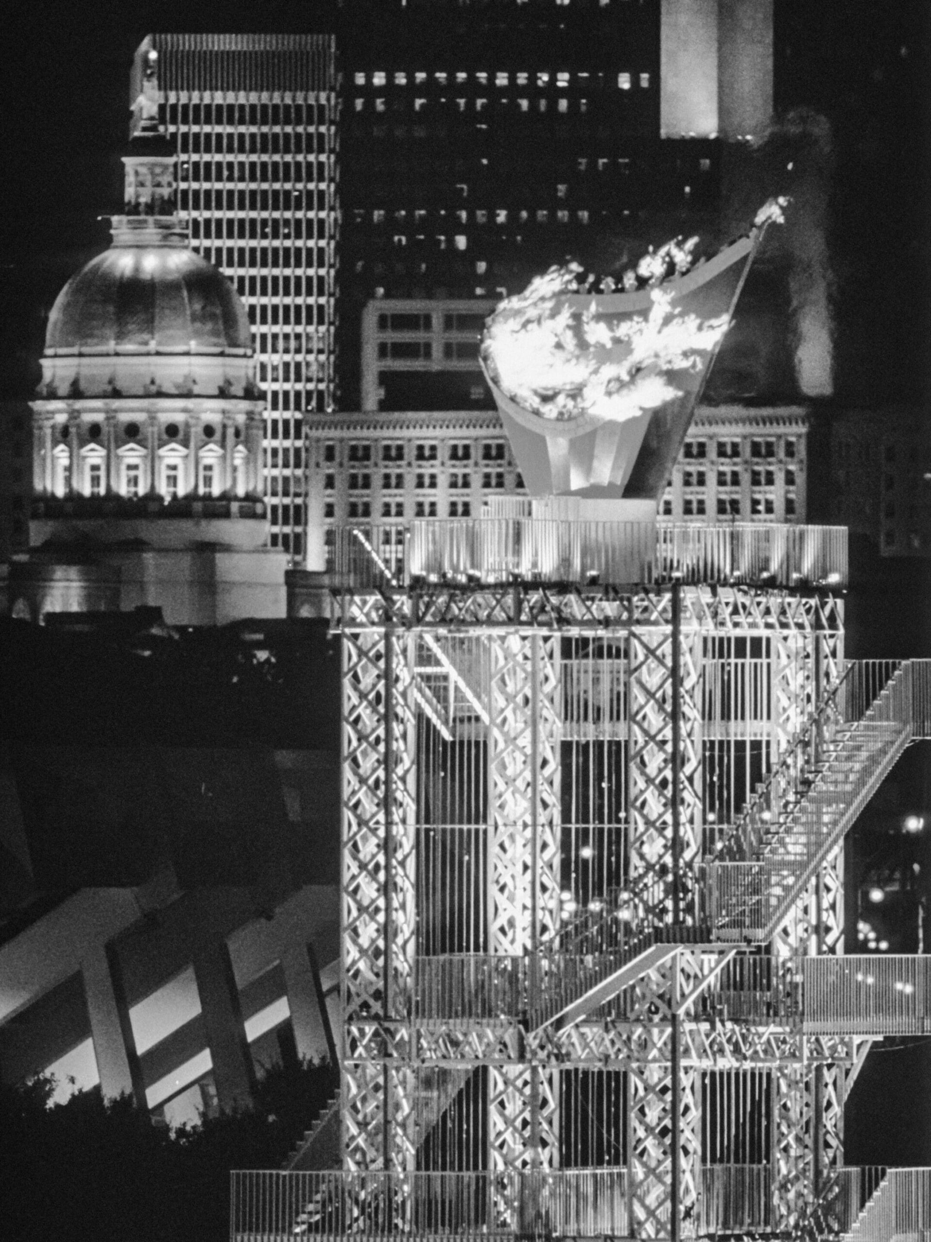 Black and white image of a lit Olympic cauldron on a metal structure, set against a cityscape with Type I Meeting Rooms and a domed building in the background.