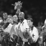 A basketball team poses together in a meeting room, proudly wearing matching uniforms and holding bouquets and medals.