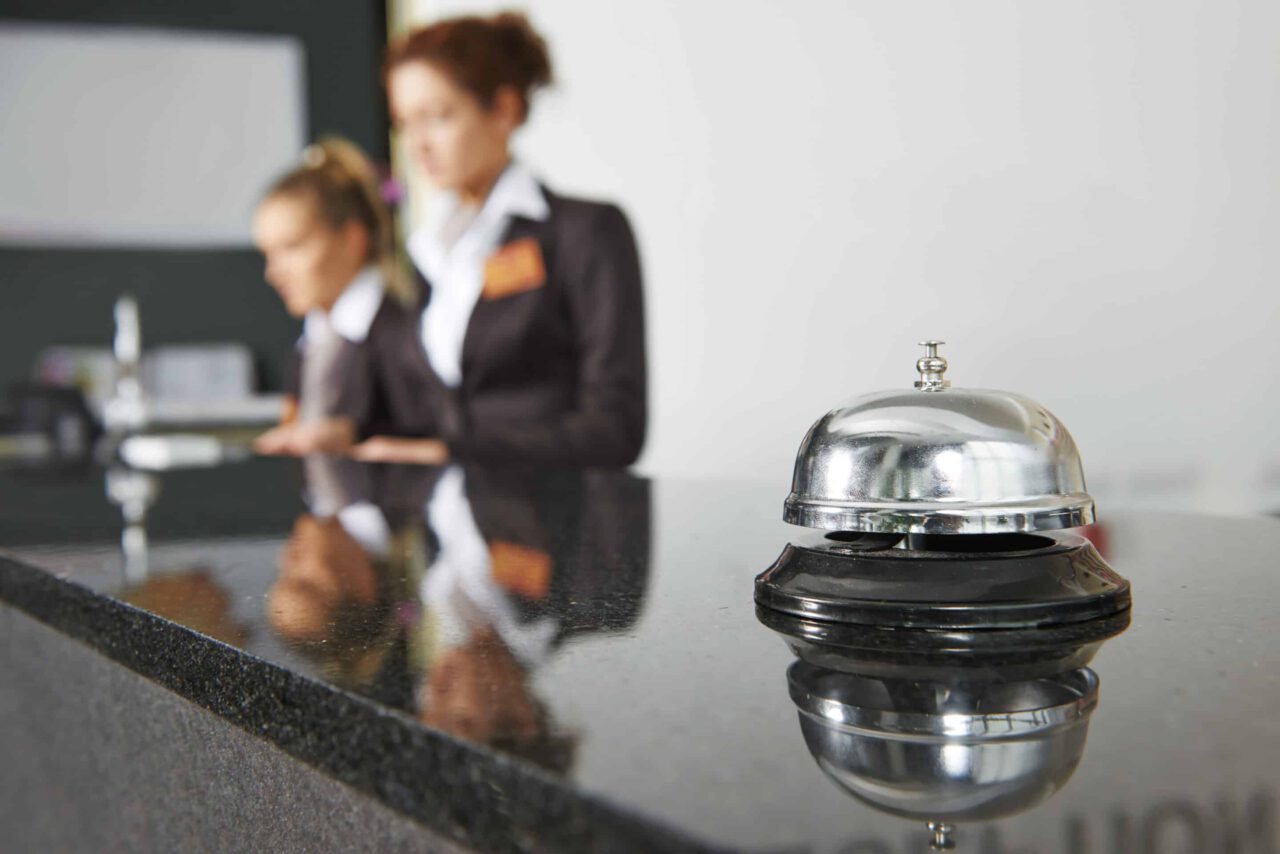 A service bell is in focus on a hotel reception desk, with two people working in the background.