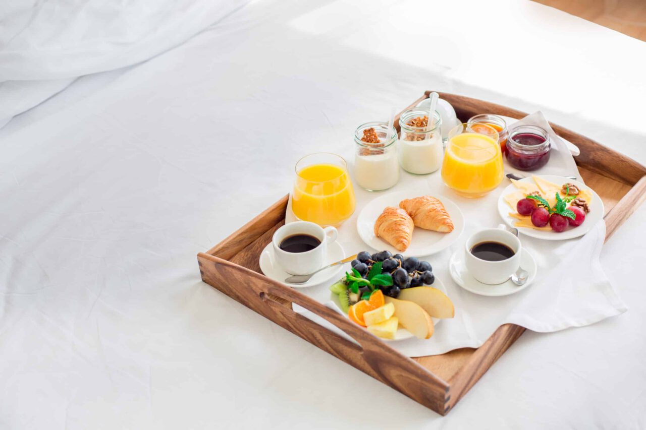 A breakfast tray on a bed with coffee, juice, croissants, fruit, yogurt, and granola.