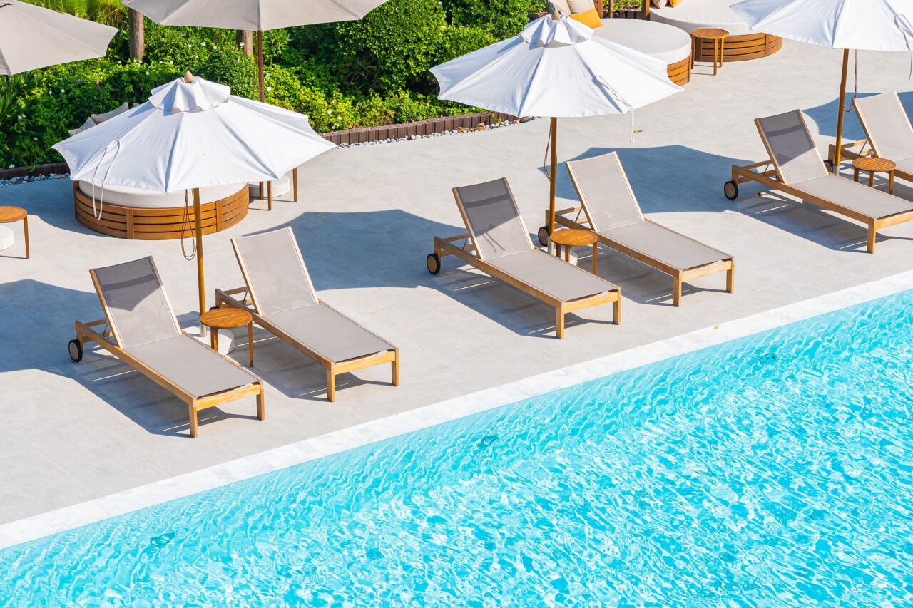 Four beige lounge chairs with white umbrellas are arranged by a bright blue swimming pool on a sunny day.