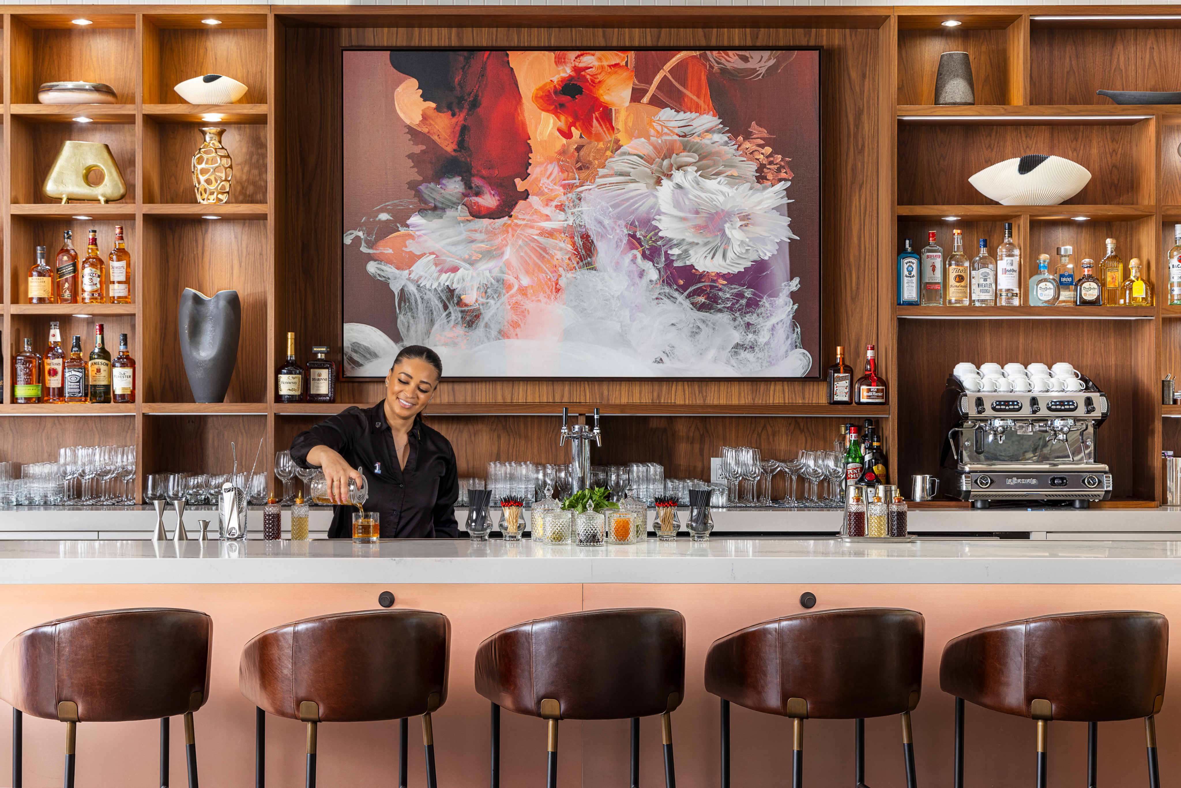 Bartender preparing a drink behind a modern bar with brown leather stools, decorative shelves, and a colorful floral artwork on the wall.