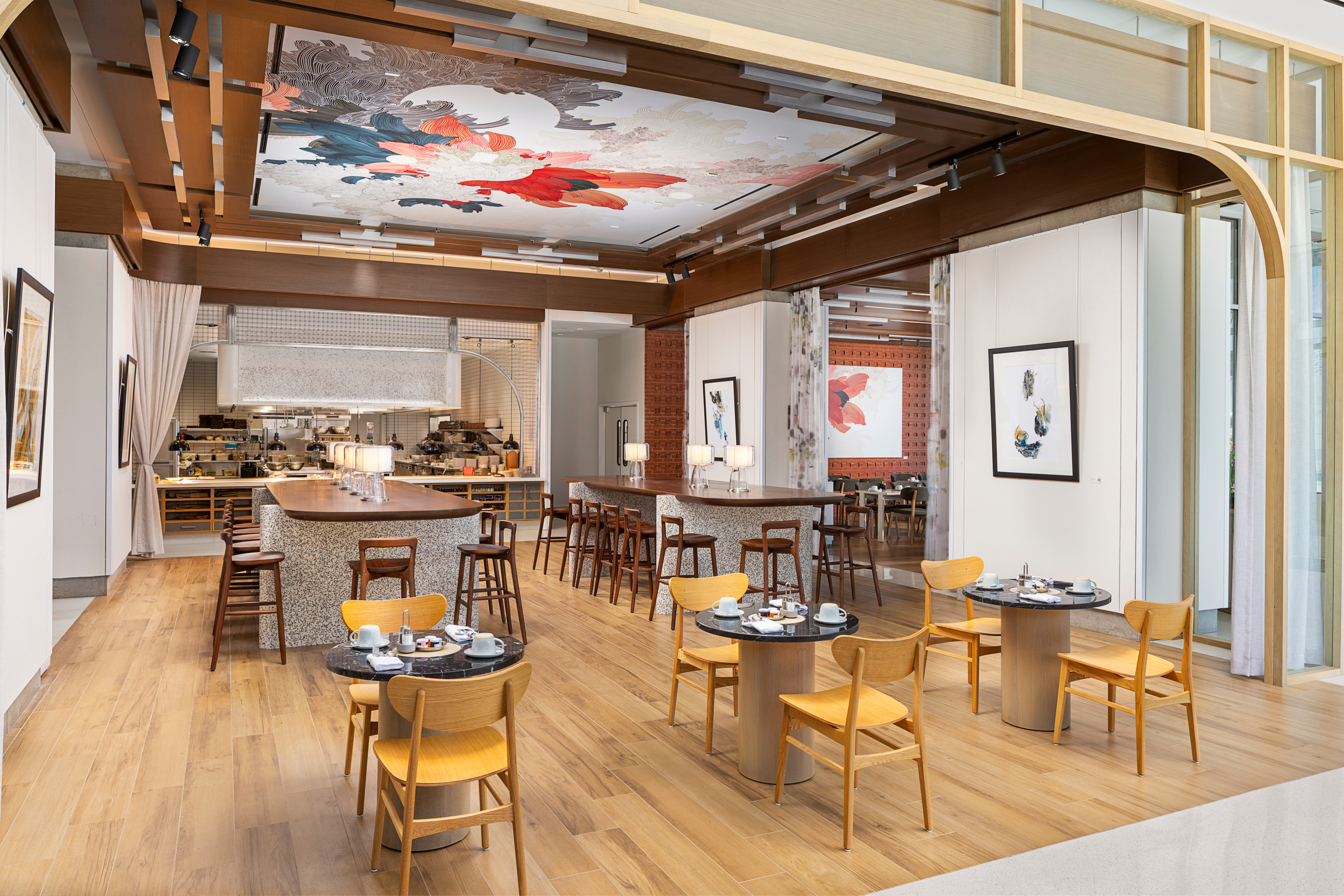 Modern restaurant interior with wooden tables and chairs, featuring a large decorative ceiling artwork and an open kitchen in the background.
