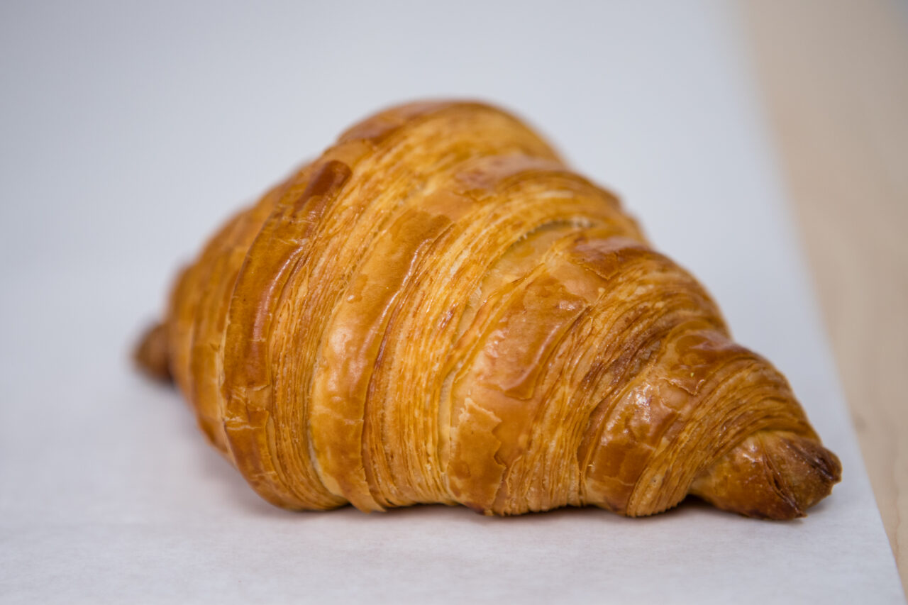 A single golden-brown croissant sits on a sheet of white parchment paper against a neutral background, evoking the warm, inviting atmosphere of Friendship Market.