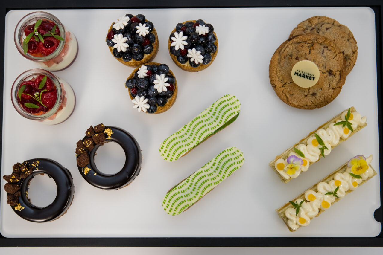 A delightful variety of desserts from Friendship Market is displayed on a white tray, including berry tarts, chocolate donuts, cookies, two green-striped eclairs, two fruit-topped parfaits, and two rectangular cream pastries.