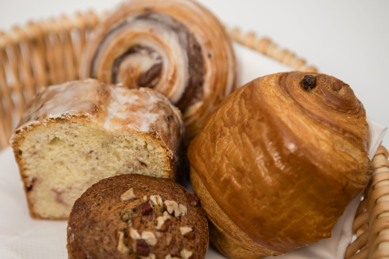 A wicker basket from Friendship Market contains a cinnamon roll, a glazed loaf slice, a pecan muffin, and a pain au chocolat on a white napkin.