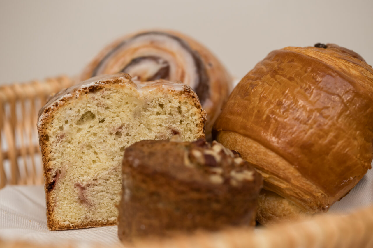 A wicker basket from Friendship Market holds assorted pastries, including a sliced loaf cake, a chocolate swirl pastry, a muffin, and a pain au chocolat on a white napkin.