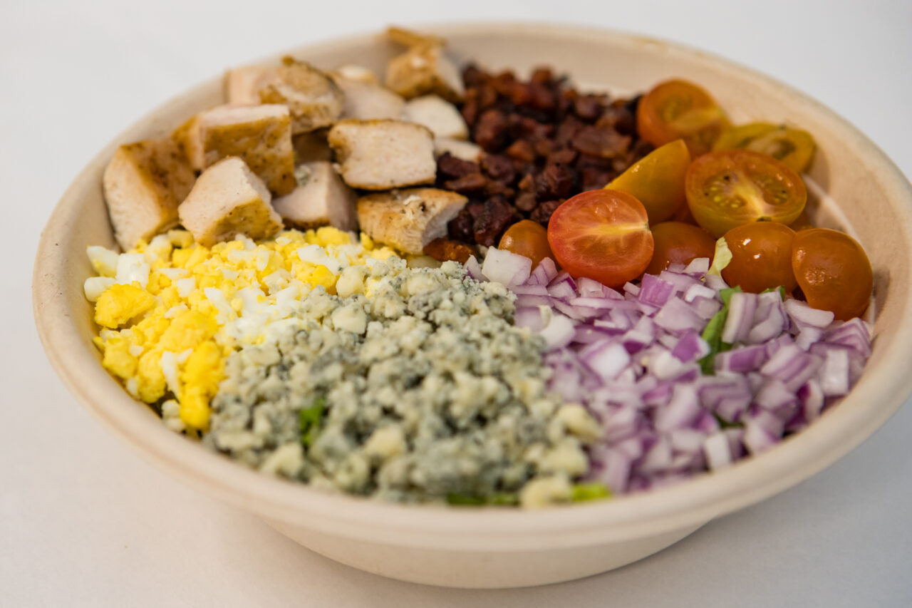A salad bowl from Friendship Market with rows of diced chicken, crumbled bacon, halved cherry tomatoes, chopped egg, blue cheese, and chopped red onion on a white surface.