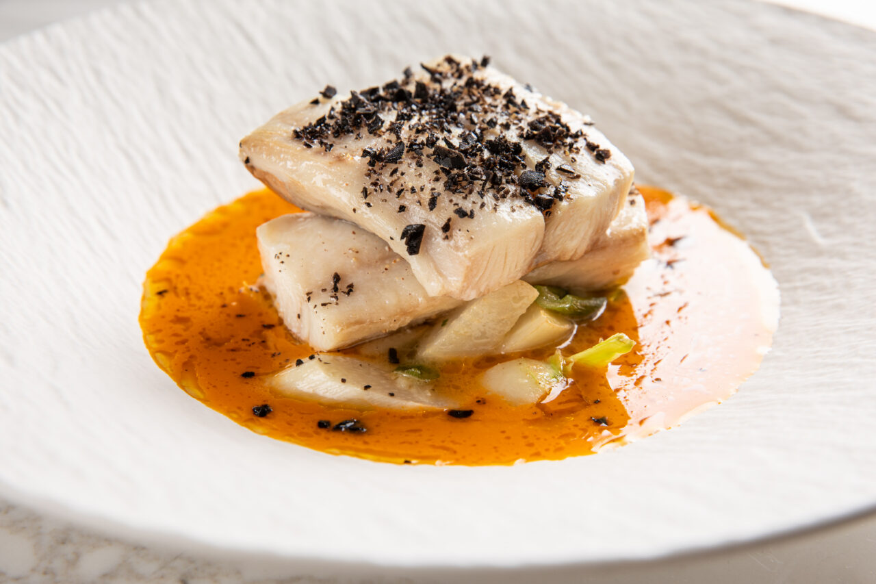Two pieces of white fish topped with black seasoning, served on vegetables with an orange sauce in a white textured bowl.