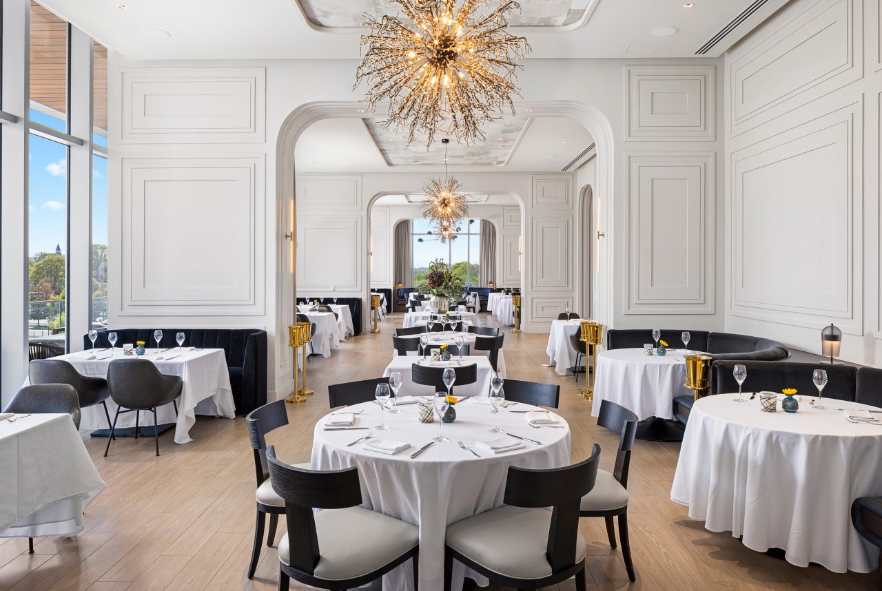 Elegant dining room with round tables covered in white cloths, black chairs, large chandeliers, and tall windows. Tables are set with glasses and napkins. Bright, spacious ambiance.