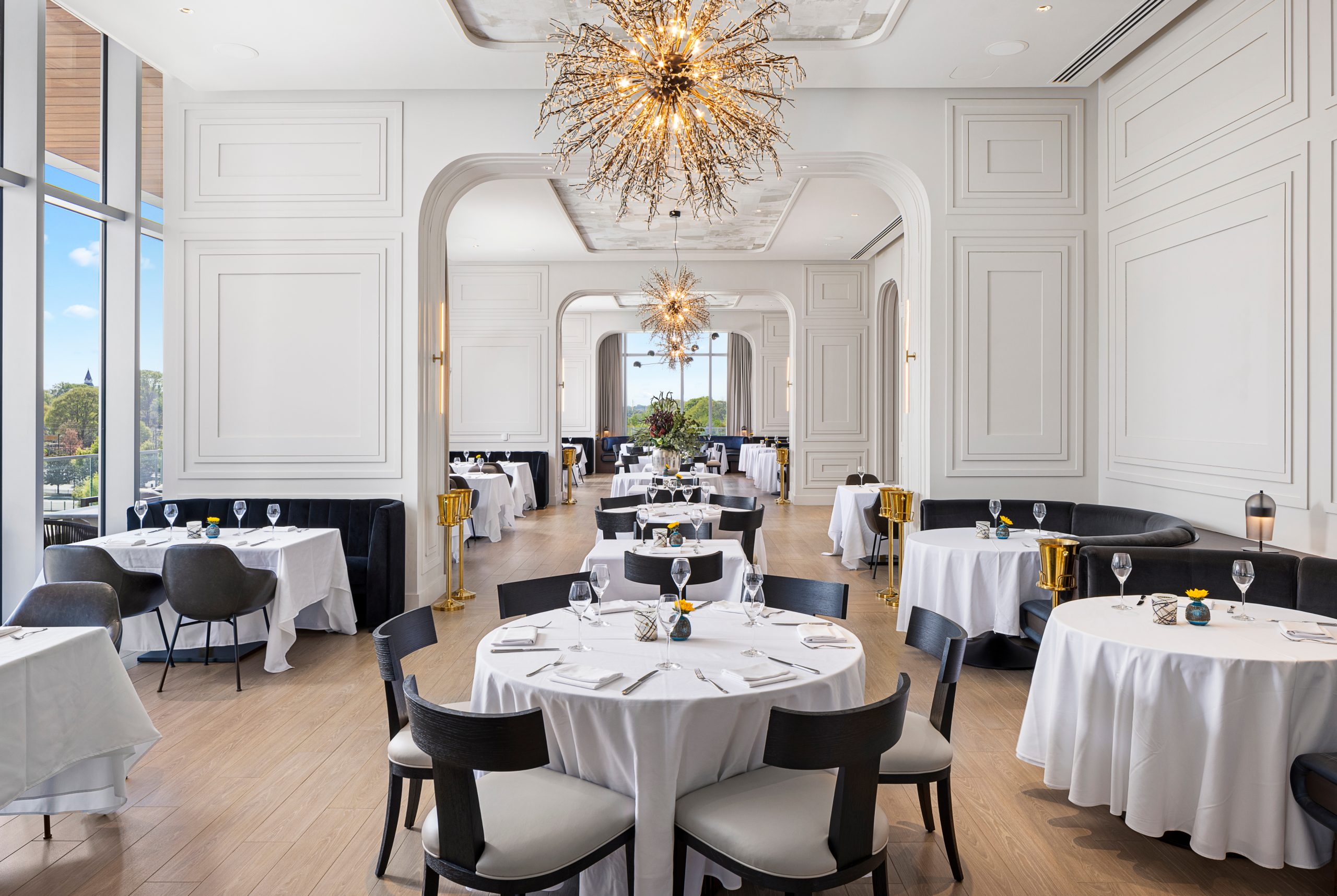 Elegant dining room with round tables covered in white cloths, black chairs, large chandeliers, and tall windows. Tables are set with glasses and napkins. Bright, spacious ambiance.