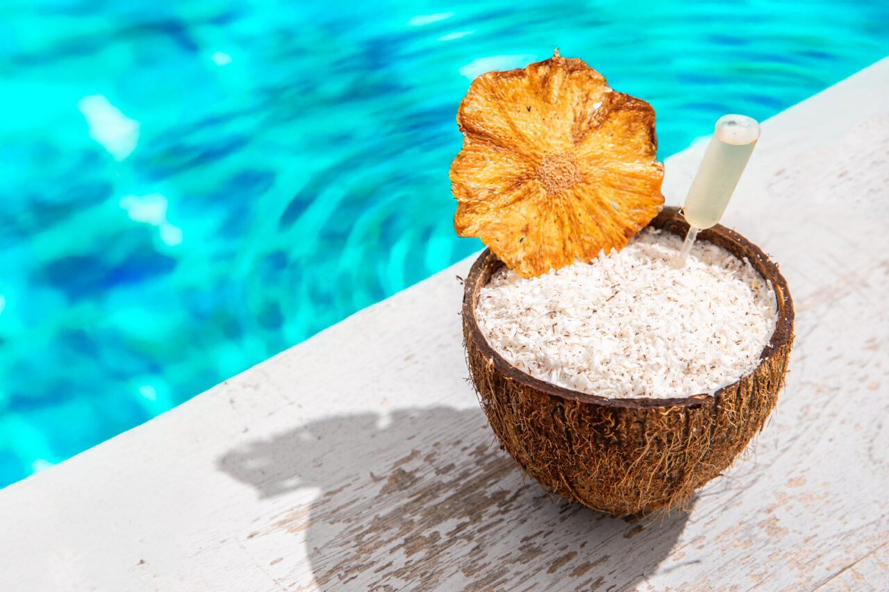 A tropical drink served in a coconut shell with a dried pineapple slice and a pipette, placed on a white ledge beside a blue swimming pool.