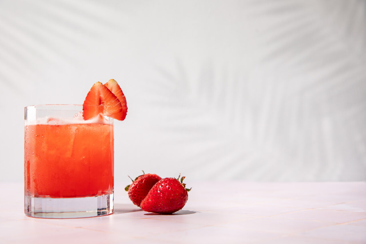 A glass of iced strawberry drink with a strawberry slice on the rim sits beside two fresh strawberries on a light pink surface.