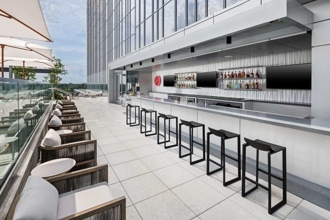 Outdoor rooftop bar with a modern design featuring high stools, a sleek counter with drinks displayed, and a row of cushioned chairs under umbrellas along a glass railing.