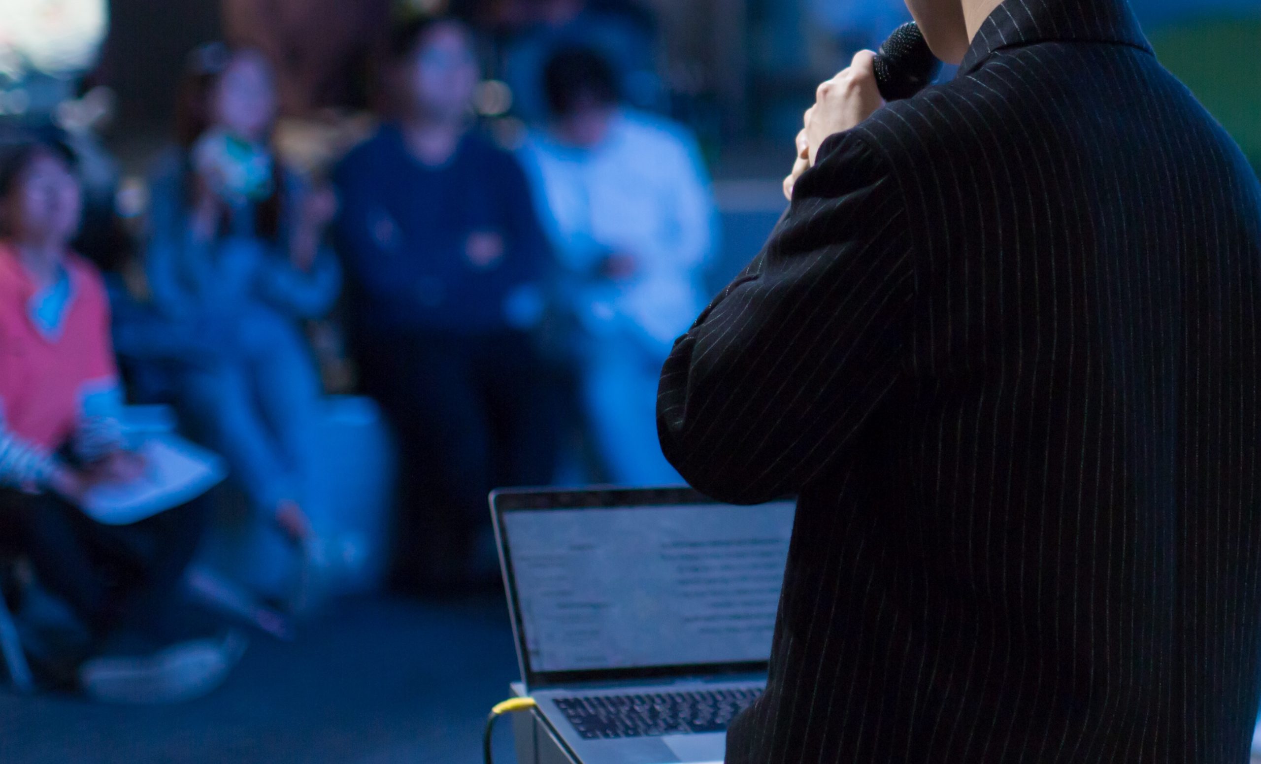 A person in a dark suit speaks into a microphone, facing a blurred audience. A laptop with an open document is on a table.