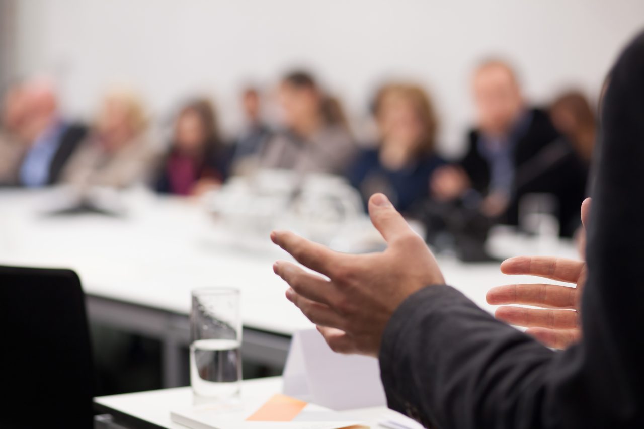 Person gesturing during a meeting in a conference room, focus on hands, with blurred attendees seated in the background.