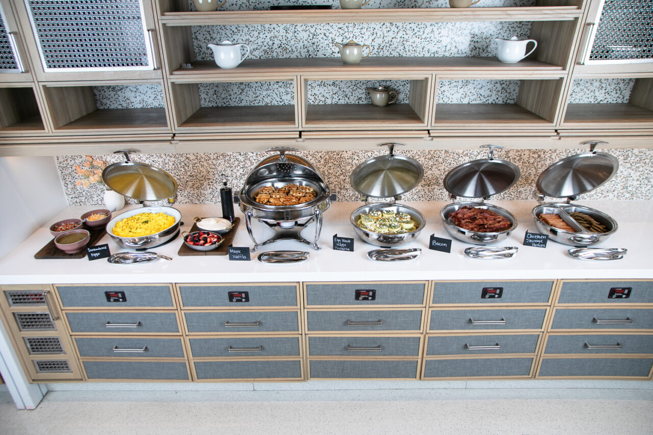 A breakfast buffet with chafing dishes containing eggs, sausages, potatoes, vegetables, and condiments on a counter with labeled signs and plates.