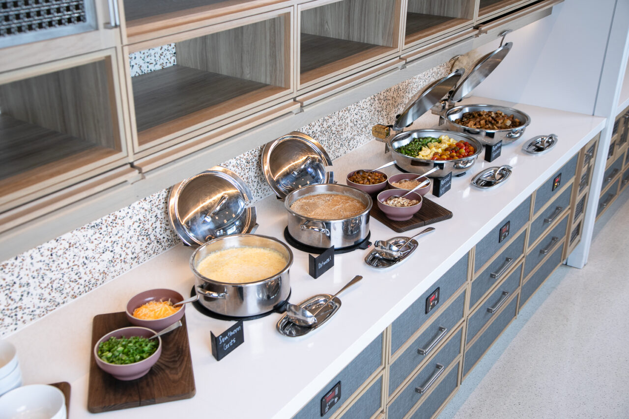 Buffet-style food setup with covered metal pots, serving utensils, bowls of toppings, and various prepared dishes on a white countertop.