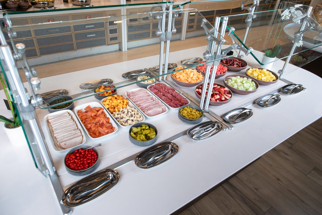 Buffet counter with various sliced meats, cheeses, and bowls of cut fruits and vegetables arranged in rows behind a glass barrier.