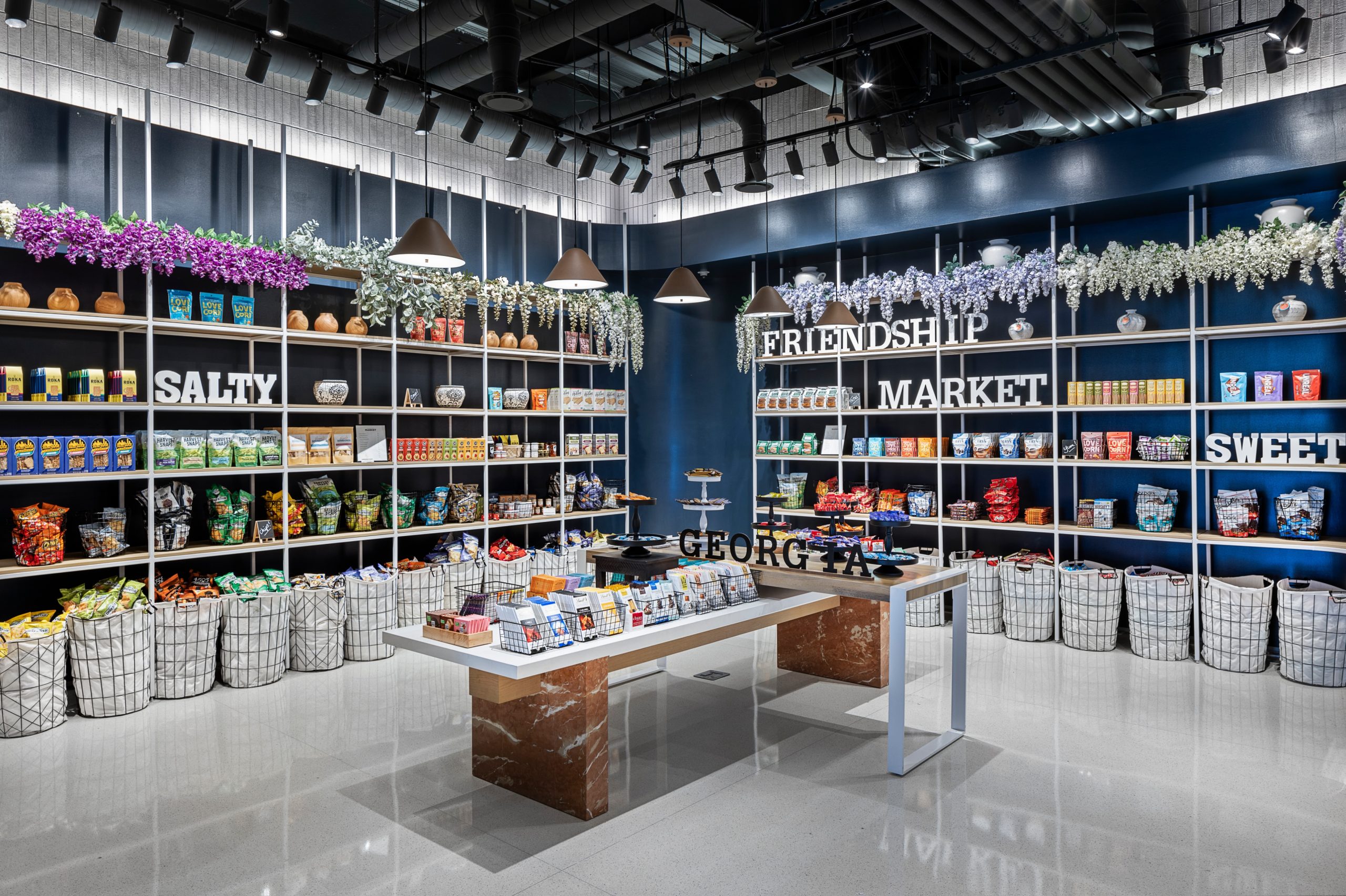 A well-organized snack shop reminiscent of a bustling market, with sections labeled "Salty" and "Sweet." Shelves display various packaged snacks, and bins hold loose items. Flowers hang above the shelves like a nod to friendship, while the floor is polished.