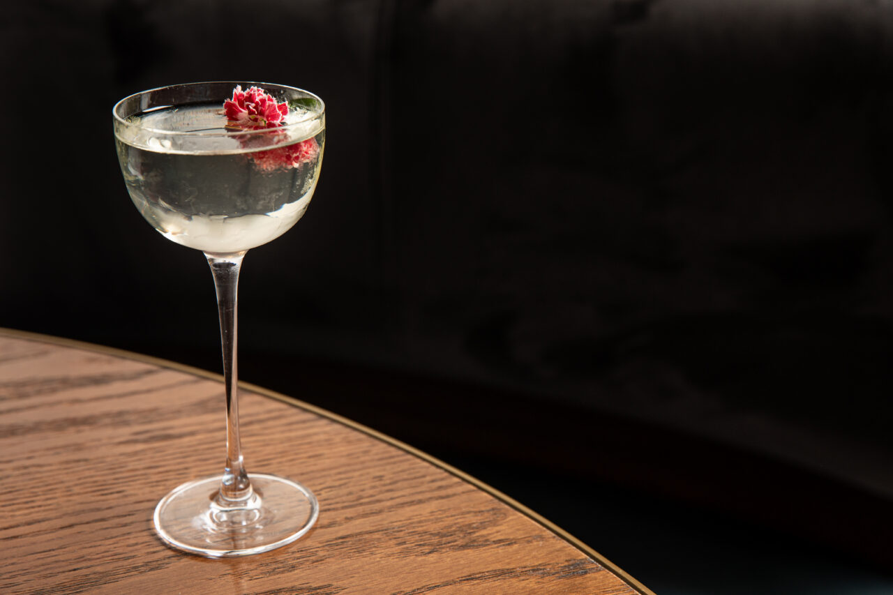 A clear cocktail in a stemmed glass garnished with a small red flower sits on a wooden table against a dark background.