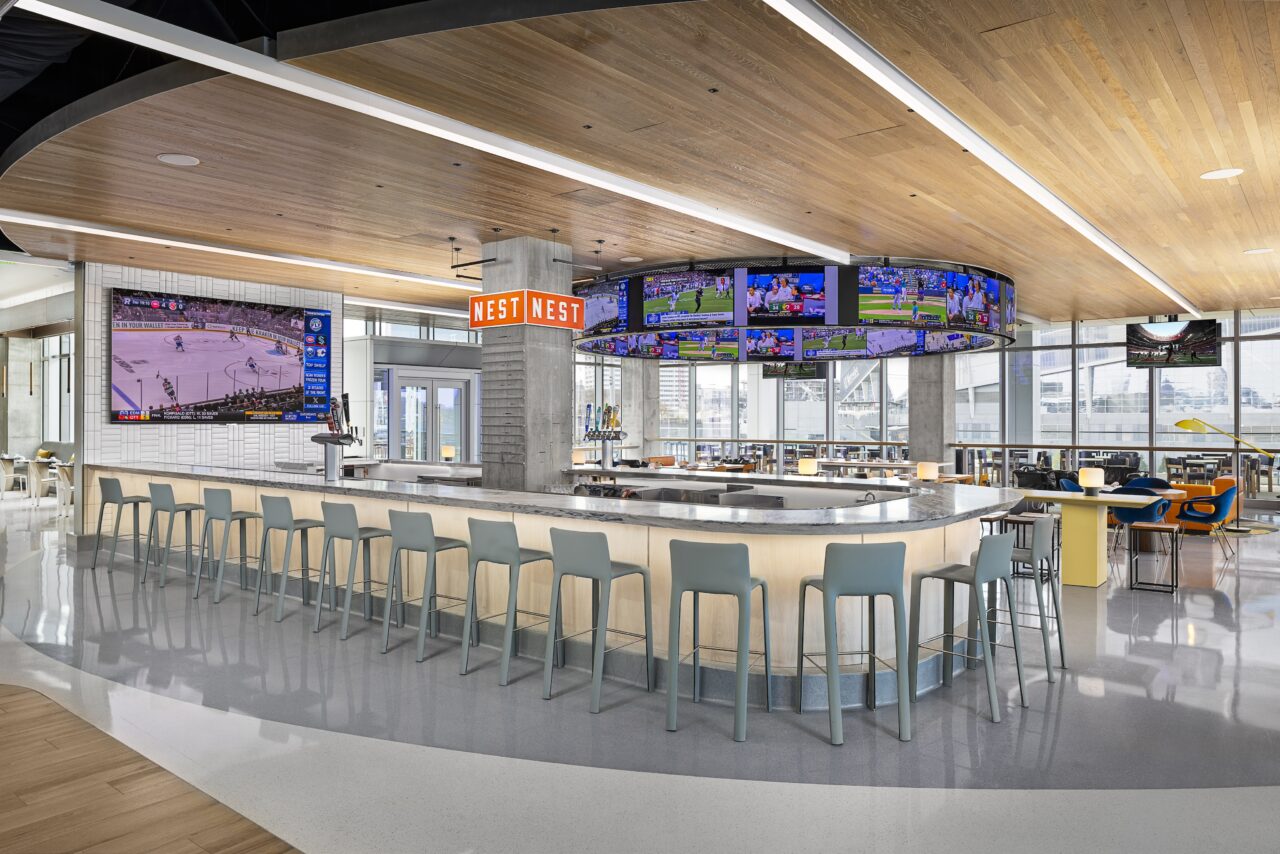 Nest on Four: A modern sports bar with a central island bar, surrounded by high stools. Multiple TVs broadcast various sports games in this bright, open space with wooden ceiling accents and large windows. It feels like a cozy nest, making everyone feel right at home.