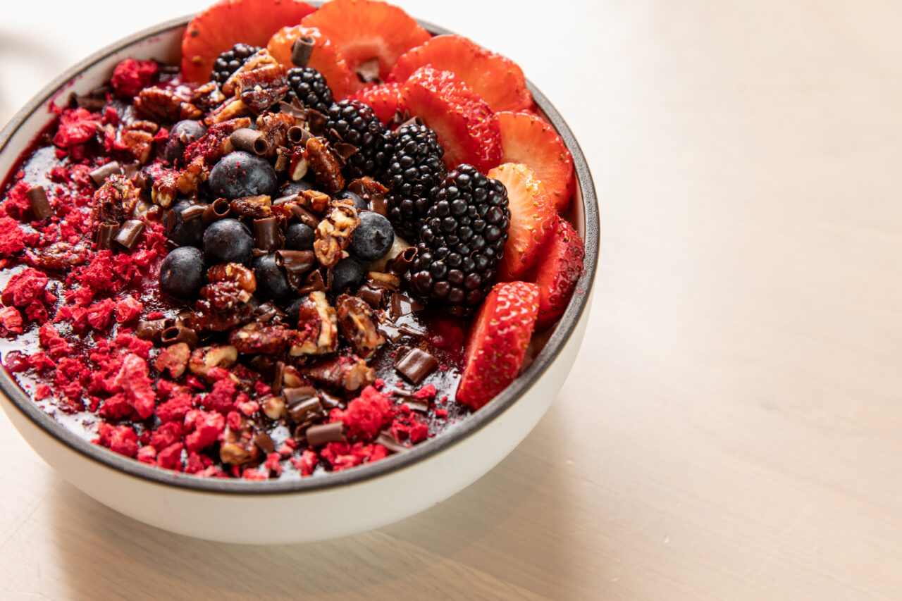 A bowl of smoothie topped with sliced strawberries, blackberries, blueberries, chopped nuts, chocolate shavings, and red berry crumbles on a light wooden surface.