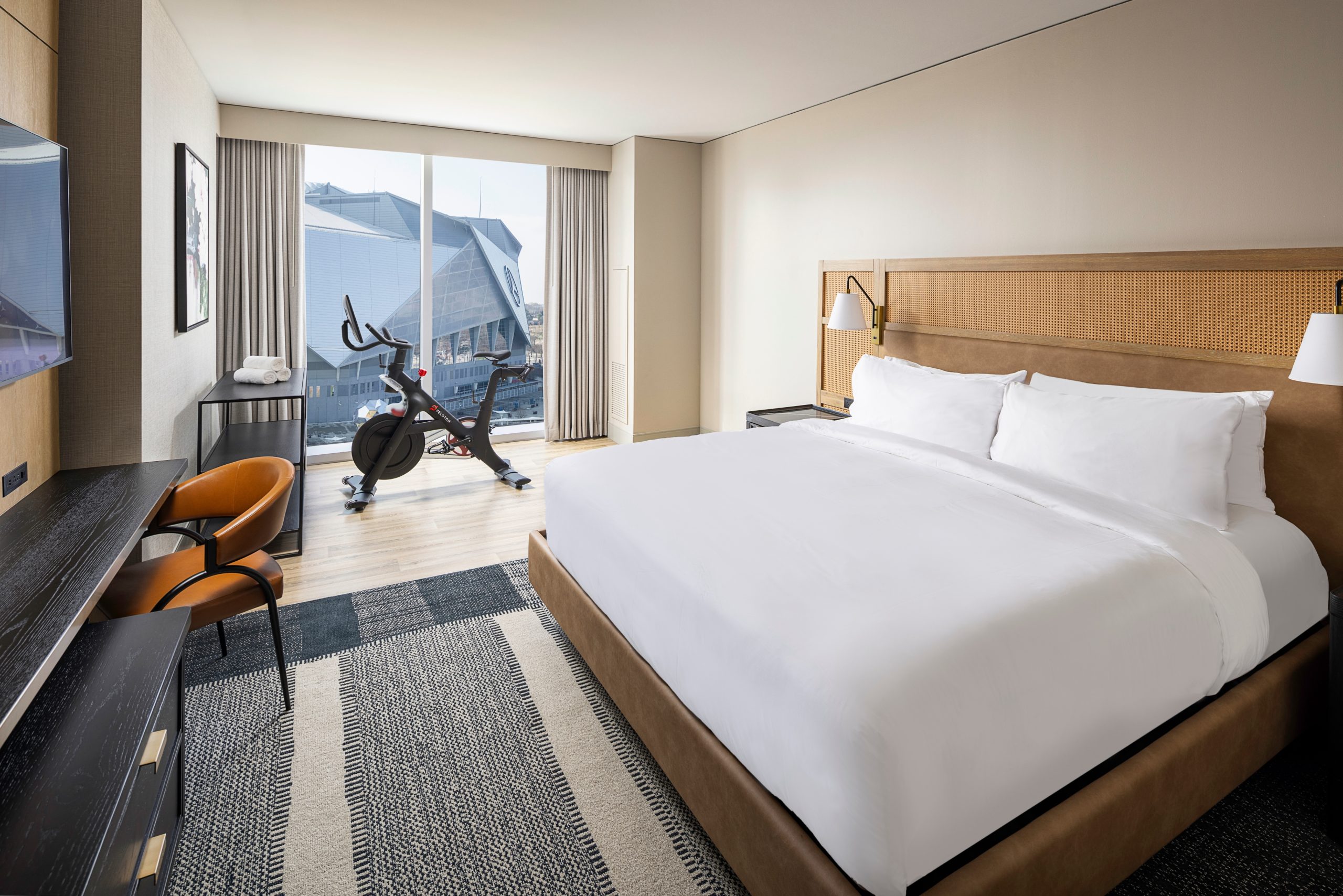 Hotel room with a large bed, desk, chair, and stationary exercise bike near a window with a view of a building.