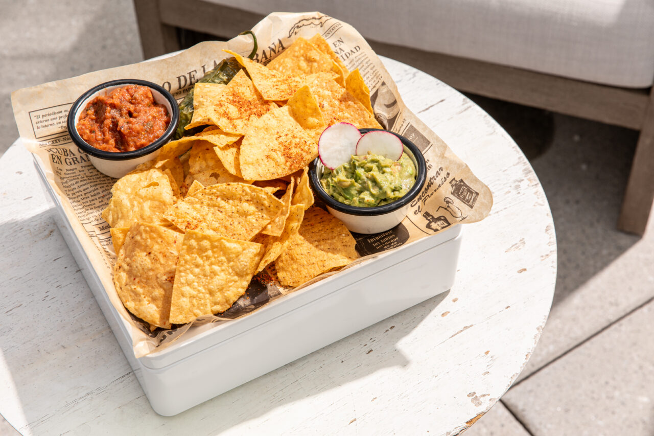 A tray with tortilla chips, a bowl of salsa, and a bowl of guacamole topped with radish slices, served on newspaper-style paper on a white table.