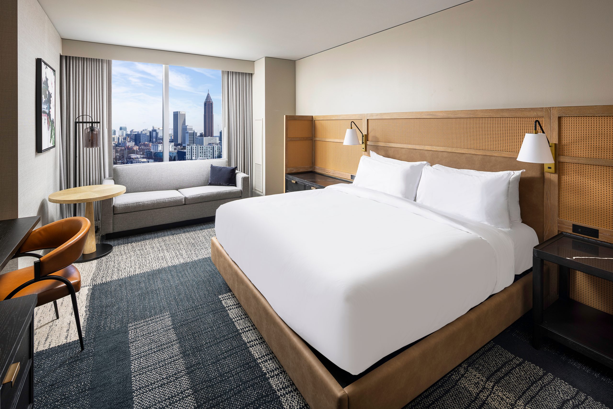 Modern hotel room featuring a large bed, wall-mounted lamps, and a sofa by the window with a city skyline view. This serene space includes wellness amenities, a round table with two chairs, and neutral decor for elevated comfort.