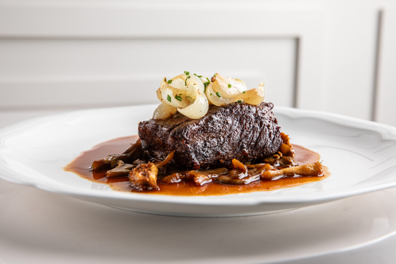 A piece of braised beef sits on a white plate, topped with pearl onions and chives, and surrounded by brown sauce with mushrooms.