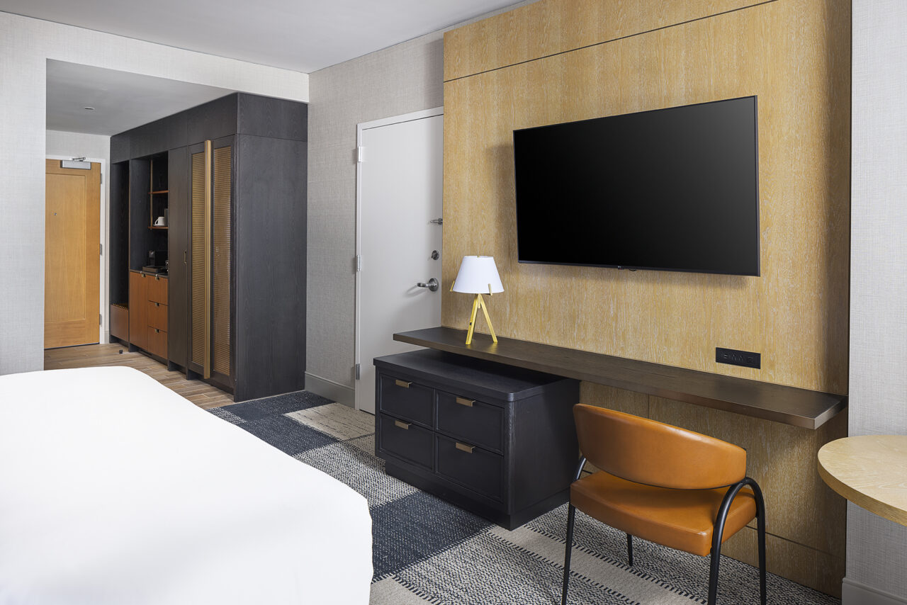 Modern hotel room with a wall-mounted TV, desk, brown chair, dresser, lamp, and a bed partially visible in the foreground. Closet and entryway in the background.