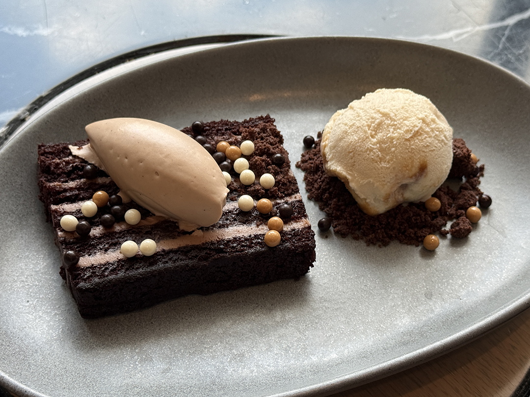 A rectangular chocolate cake slice with chocolate mousse and crisp pearls, served with vanilla ice cream on chocolate crumbs, elegantly presented at Nest on Four on a gray oval plate.