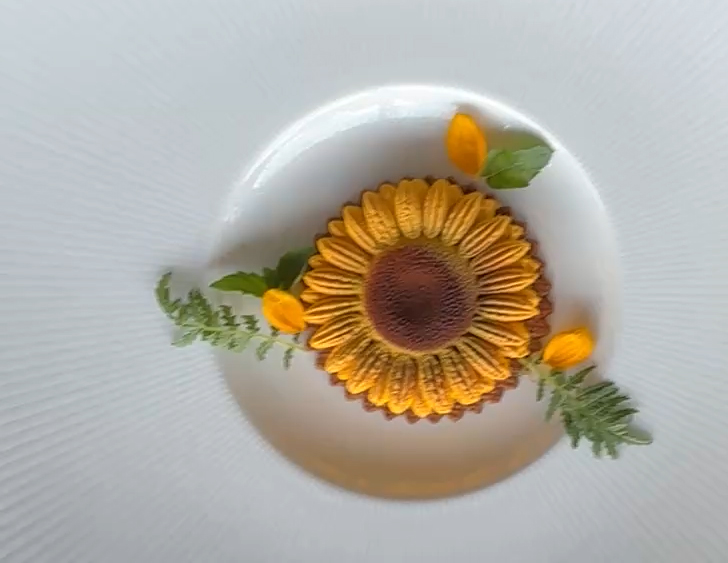 A dessert arranged to resemble a sunflower, with yellow petals, a brown center, green leaves, and yellow accents on a white plate.