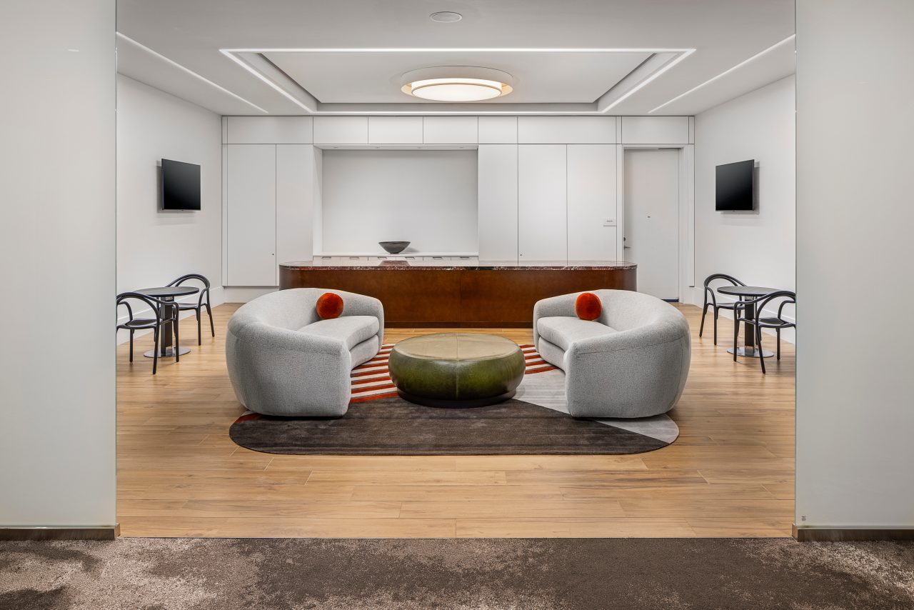 A chic Heritage Lounge features two gray armchairs, a green ottoman, and a striped rug. The backdrop boasts a minimalist kitchen with two small dining tables and chairs, merging contemporary comfort with timeless elegance.