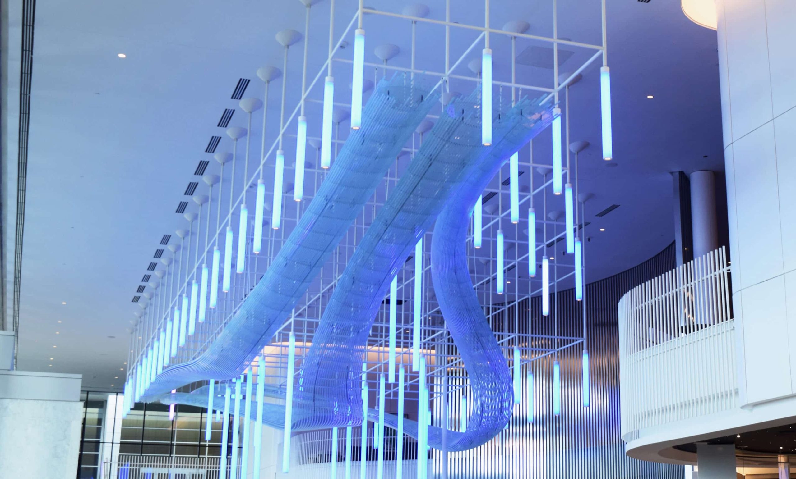 Large abstract sculpture with blue lighting hangs from the ceiling in a modern building. The structure includes vertical rods and curved elements, creating a flowing, dynamic appearance.