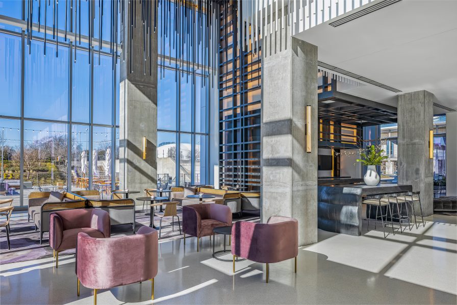 Modern lounge with velvet chairs, concrete columns, and large windows. Sunlit space features a bar with high stools and ceiling decor with vertical black lines.