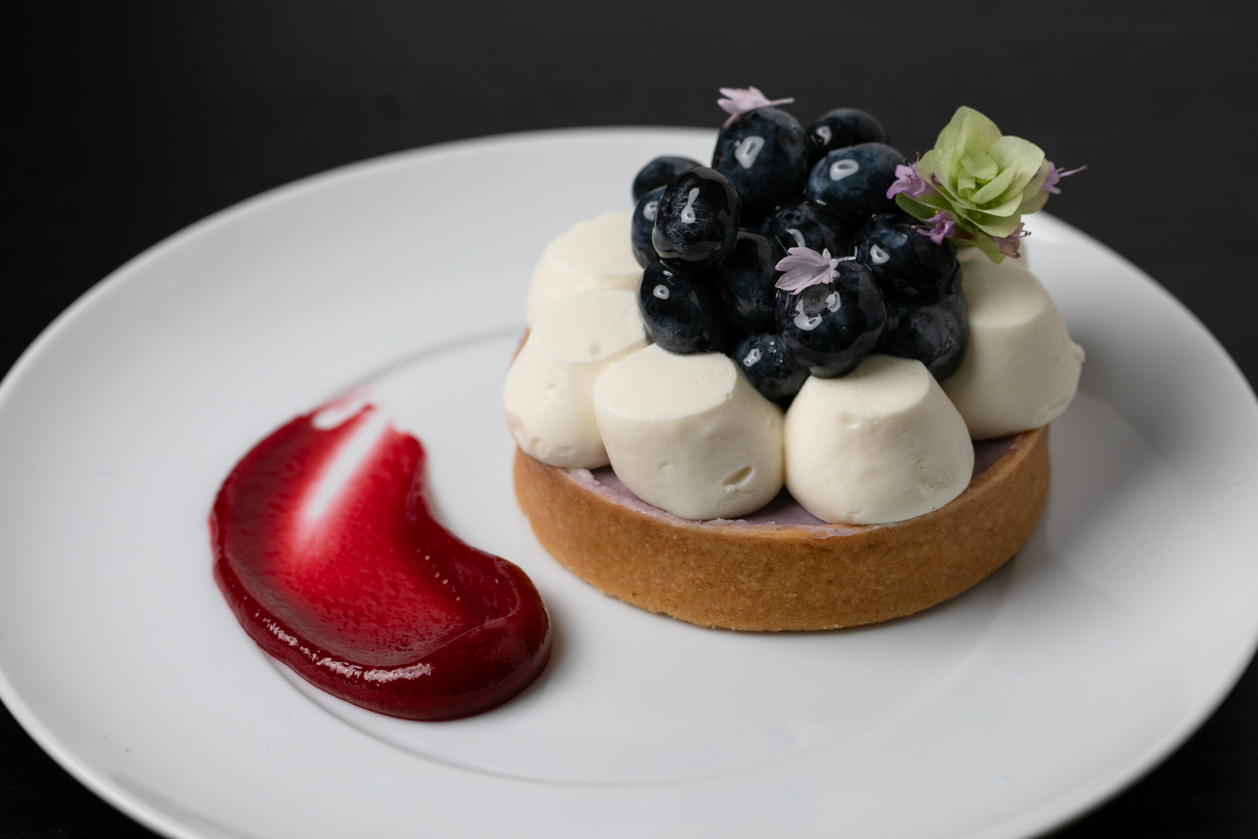 A tart topped with white meringue, blueberries, and small flowers sits on a white plate with a smear of red berry sauce.