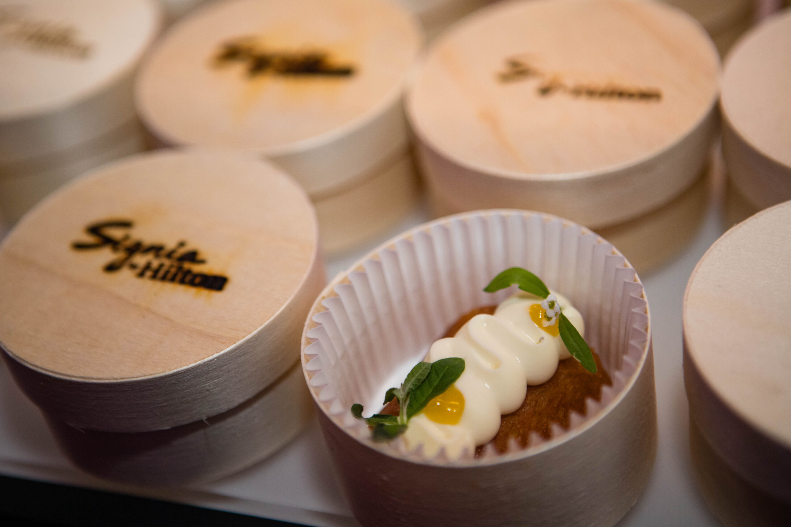 A small dessert, perfect for a private dining experience, is garnished with cream and leaves in a paper liner, surrounded by wooden containers featuring the "Signia by Hilton" branding.